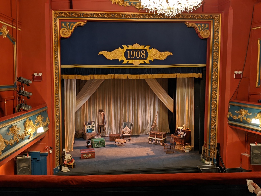 Image of the proscenium stage at Todmorden Hippodrome taken from the circle. The stage is simply set for the 2022 production of 'Little Women' with a writing desk, trunks, a doll's house, and chairs.